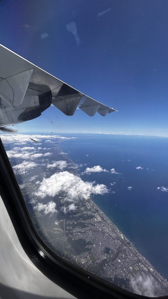 トキエアの窓から見た翼と海岸線。青い海と白い雲、海沿いの街が見える