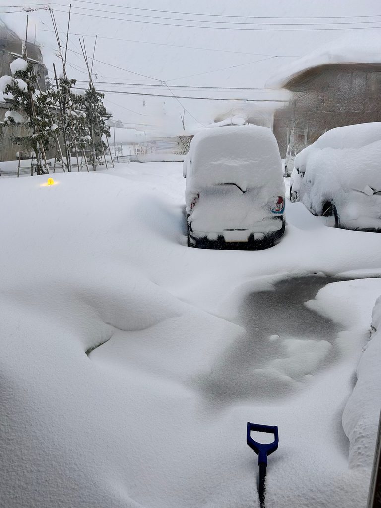 大雪の駐車場。雪に埋もれた車の手前に、雪かき用のスコップが立っている